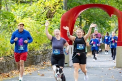 Two women wave while other racers run near the iconic red arch sculpture  at the Artful Dash 5K Run/Walk on the Karl Stirner Arts Trail in Easton, Pennsylvania.