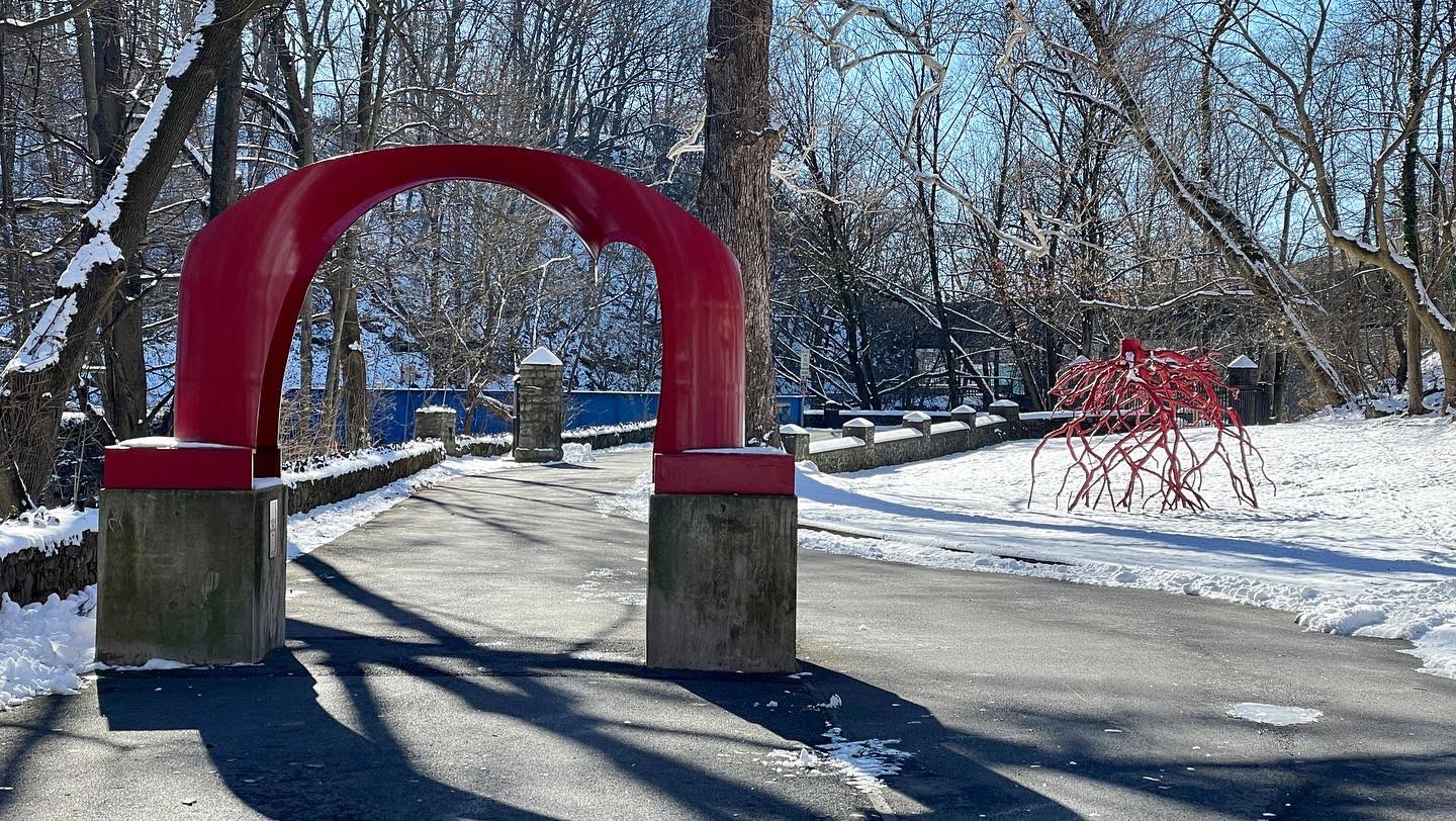 The iconic red arch sculpture with snow-covered ground and the Late Bronze Root sculpture in the distance on the Karl Stirner Arts Trail