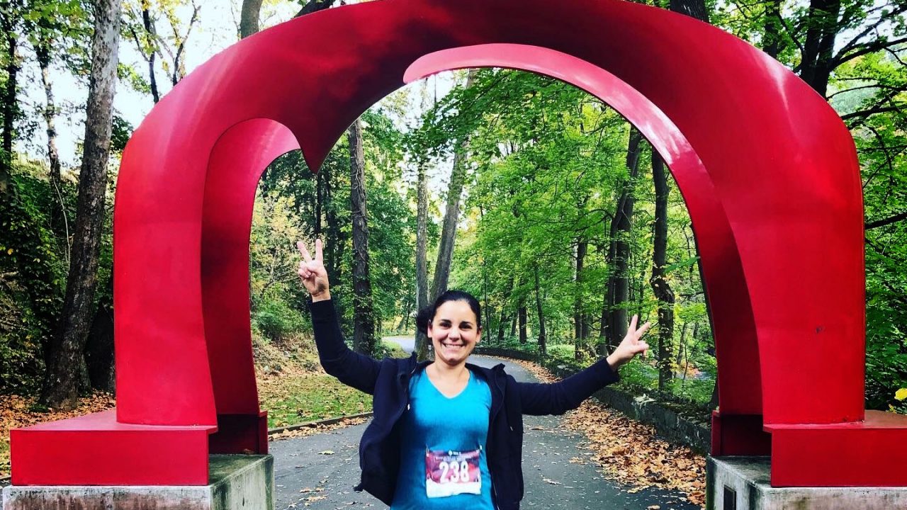 A runner smiles and gives the peace sign with both hands while standing under the red arch sculpture on Hanging pieces of art made from fabric on the Karl Stirner Arts Trail in Easton, Pennsylvania.