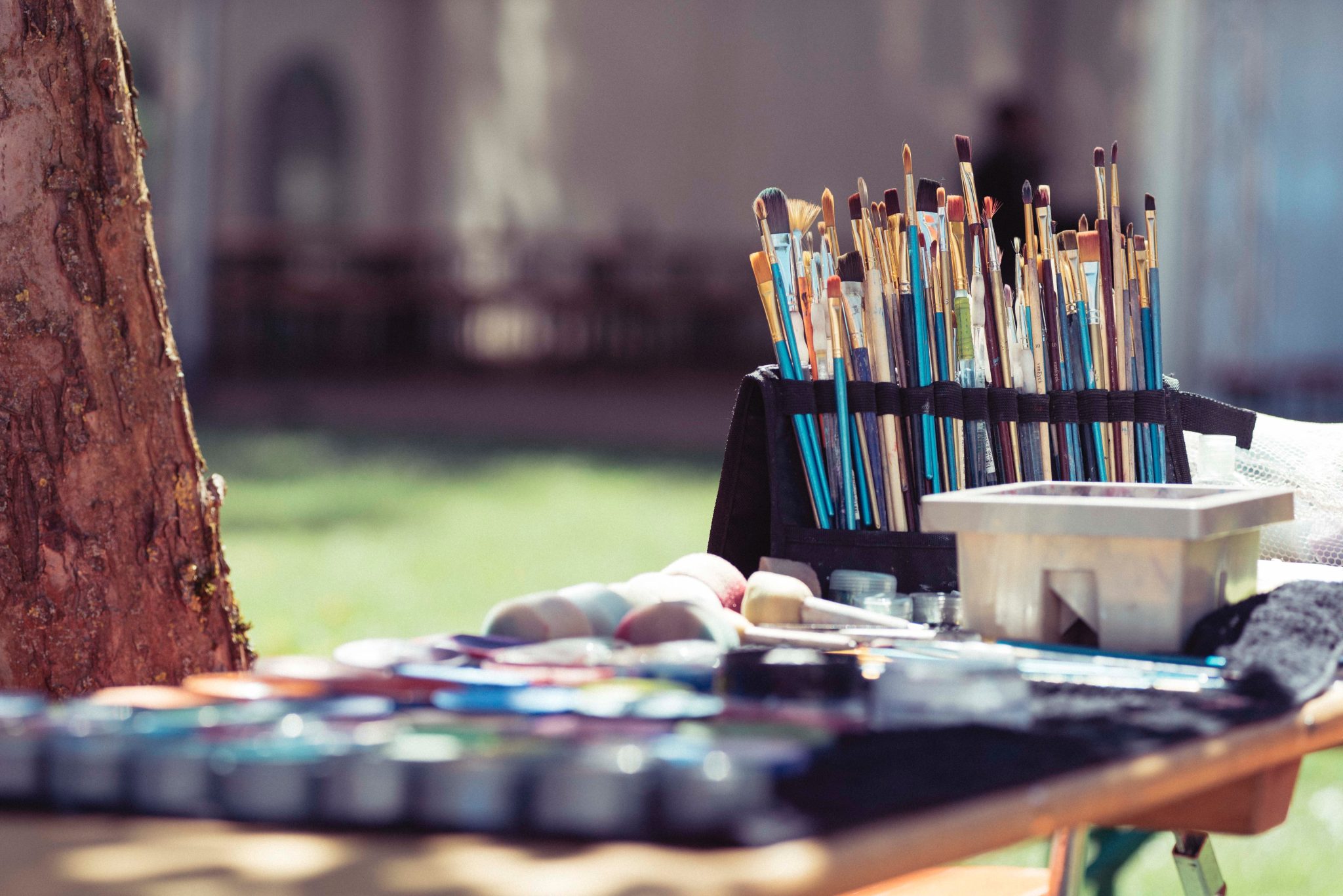 A set of paint brushes and paints on a table near a tree outdoors