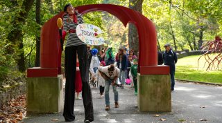A woman on stilts stands by the red arch sculpture as children in costumes with their parents are behind the arch during the Come as You Art event on the Karl Stirner Arts Trail in Easton, Pennsylvania.