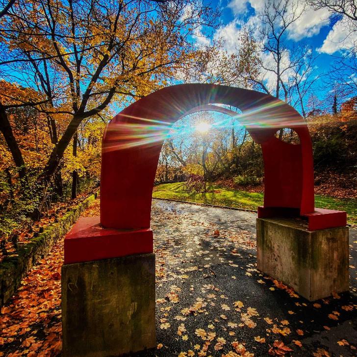 Sunlight shines from behind an autumn tree and below the top curve of the iconic red arch sculpture on the Karl Stirner Arts Trail in Easton, Pennsylvania.