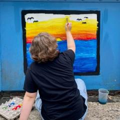 A boy paints a yellow and red sky above blue water with birds flying above on the Young Masters Wall at the Karl Stirner Arts Trail in Easton, Pennsylvania.