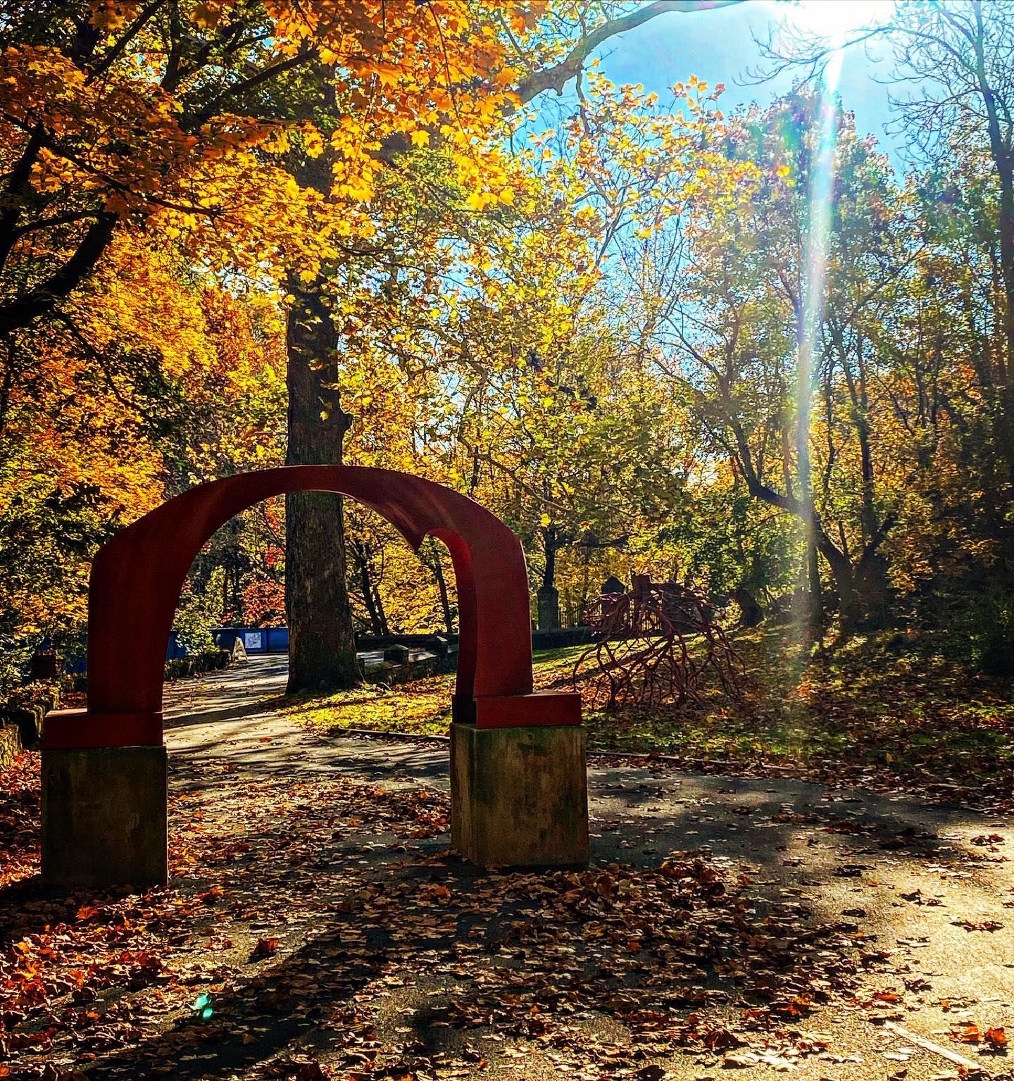 The iconic red arch sculpture and Steve Tobin's Late Bronze Root stand near trees with autumn leaves on the Karl Stirner Arts Trail in Easton, Pennsylvania.